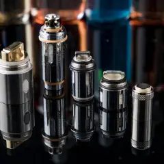 Plusieurs résistances pour cigarette électronique, certaines dorées, d'autres argentées, sont alignées sur un fond sombre avec des flacons de e-liquides flous.