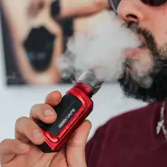 Homme barbu vapotant une cigarette électronique rouge. Il cherche à vapoter sans nicotine pour maigrir.