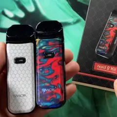 Deux vapoteuses Smok Nord 2, une blanche à motifs nid d'abeille, l'autre colorée. La boîte du kit Smok Nord 2 est visible.