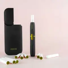 Un étui IQOS noir ouvert, un appareil de cigarette électronique qui chauffe, et plusieurs bâtonnets de tabac sur fond rose pâle.