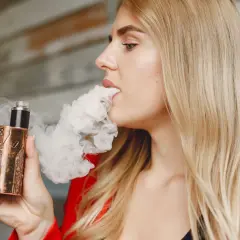 Une femme blonde vapote une cigarette électronique au design unique, exhalant un nuage de vapeur.