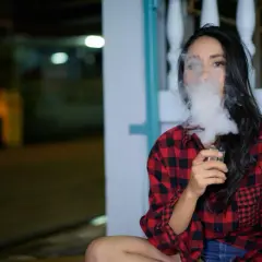 Une femme en chemise à carreaux rouges et noirs exhale un nuage de vapeur d'une cigarette électronique, son souffle se mêlant à la nuit.