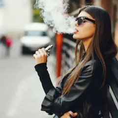 Jeune femme vapotant, lunettes de soleil, blouson en cuir. La question "est ce que la cigarette électronique fait grossir ?" reste ouverte.