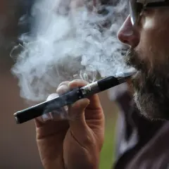 Un homme barbu fume une cigarette électronique, exhalant un nuage de vapeur. Il semble apprécier ce moment, peut-être en train de fumer et vapoter en même temps.
