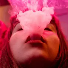 Une femme exhale de la vapeur d'une cigarette électronique, la fumée rose s'élevant autour de son visage. À gauche, une couronne rouge ornée.