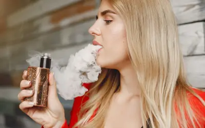 Une femme blonde vapote une cigarette électronique au design unique, exhalant un nuage de vapeur.