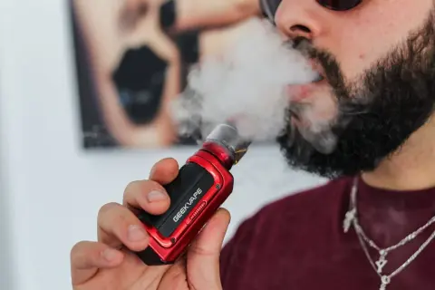 Homme barbu vapotant une cigarette électronique rouge. Il cherche à vapoter sans nicotine pour maigrir.