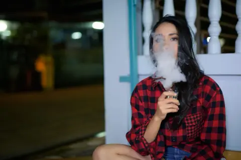 Une femme en chemise à carreaux rouges et noirs exhale un nuage de vapeur d'une cigarette électronique, son souffle se mêlant à la nuit.