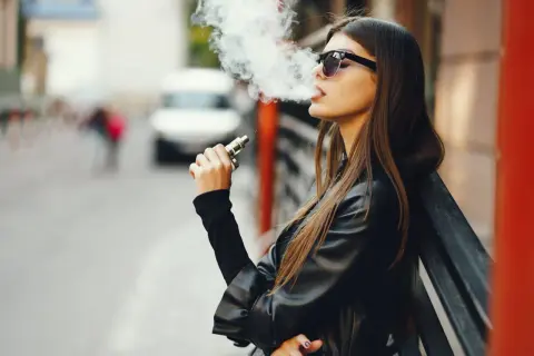 Jeune femme vapotant, lunettes de soleil, blouson en cuir. La question "est ce que la cigarette électronique fait grossir ?" reste ouverte.