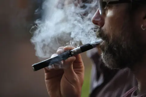 Un homme barbu fume une cigarette électronique, exhalant un nuage de vapeur. Il semble apprécier ce moment, peut-être en train de fumer et vapoter en même temps.