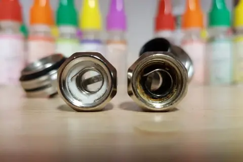 Deux résistances de cigarette électronique sur une table en bois. L'une est neuve, l'autre montre les signes d'une résistance crâmée après avoir vapoté.