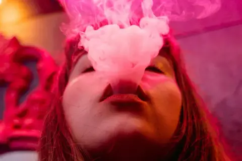 Une femme exhale de la vapeur d'une cigarette électronique, la fumée rose s'élevant autour de son visage. À gauche, une couronne rouge ornée.