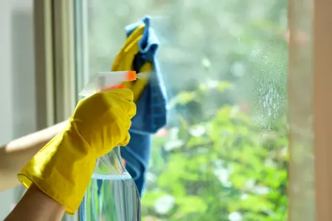 Main tenant, une main gantée de jaune pulvérise du vinaigre blanc sur une fenêtre pour nettoyer la résistance de la saleté.