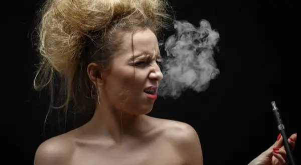 Femme aux cheveux ébouriffés, grimace en vapotant, la cigarette électronique semble lui causer une toux grasse.