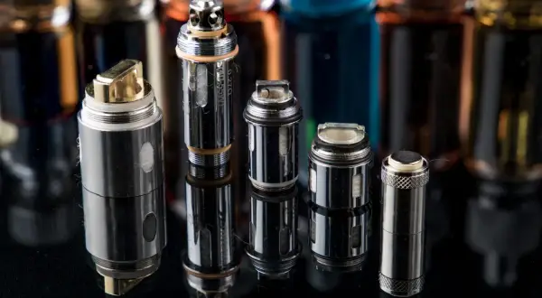 Plusieurs résistances pour cigarette électronique, certaines dorées, d'autres argentées, sont alignées sur un fond sombre avec des flacons de e-liquides flous.