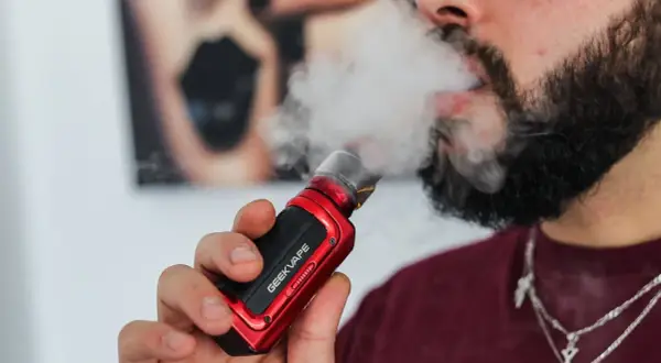 Homme barbu vapotant une cigarette électronique rouge. Il cherche à vapoter sans nicotine pour maigrir.
