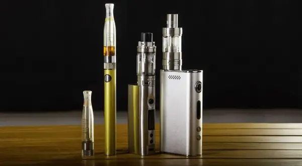 Collection de cigarettes électroniques, dont une dorée et une argentée, sur une table en bois. Ces appareils de vapotage, souvent à basse tension, offrent une alternative moderne.