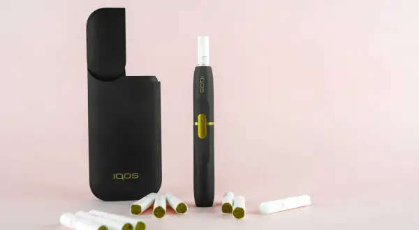 Un étui IQOS noir ouvert, un appareil de cigarette électronique qui chauffe, et plusieurs bâtonnets de tabac sur fond rose pâle.