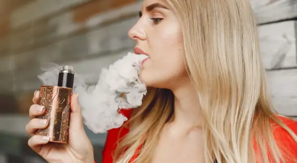 Une femme blonde vapote une cigarette électronique au design unique, exhalant un nuage de vapeur.