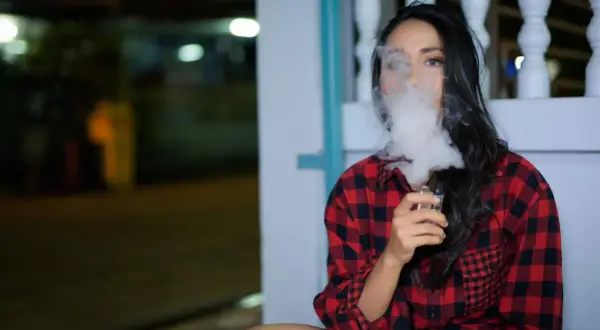 Une femme en chemise à carreaux rouges et noirs exhale un nuage de vapeur d'une cigarette électronique, son souffle se mêlant à la nuit.