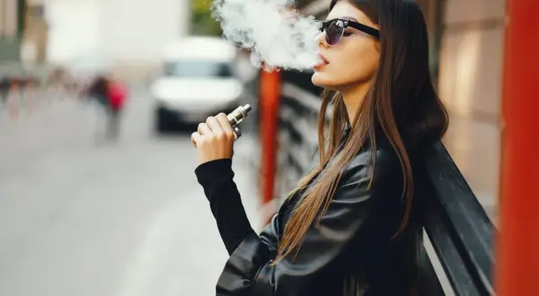 Jeune femme vapotant, lunettes de soleil, blouson en cuir. La question "est ce que la cigarette électronique fait grossir ?" reste ouverte.