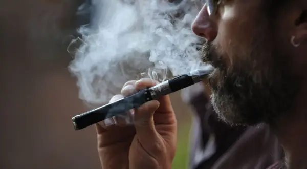 Un homme barbu fume une cigarette électronique, exhalant un nuage de vapeur. Il semble apprécier ce moment, peut-être en train de fumer et vapoter en même temps.