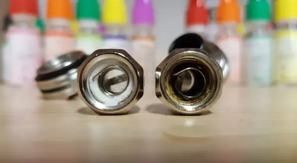 Deux résistances de cigarette électronique sur une table en bois. L'une est neuve, l'autre montre les signes d'une résistance crâmée après avoir vapoté.