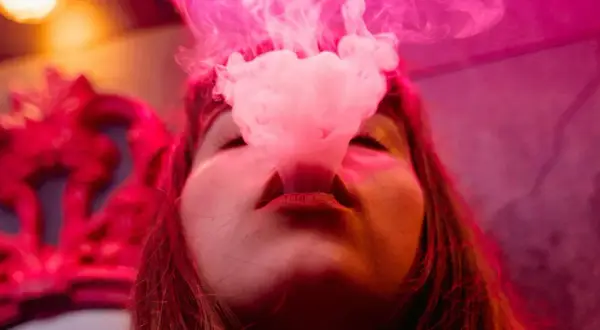 Une femme exhale de la vapeur d'une cigarette électronique, la fumée rose s'élevant autour de son visage. À gauche, une couronne rouge ornée.