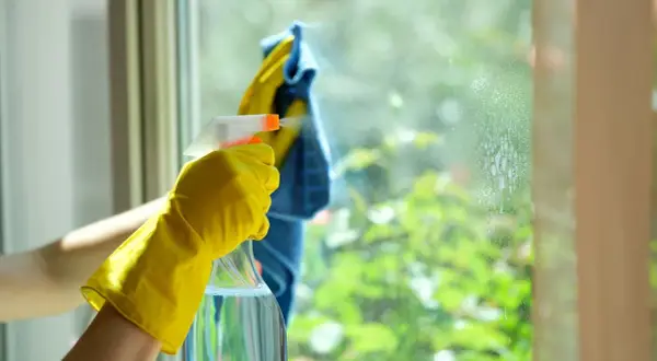 Main tenant, une main gantée de jaune pulvérise du vinaigre blanc sur une fenêtre pour nettoyer la résistance de la saleté.
