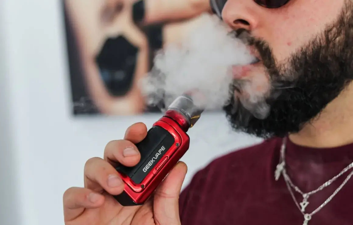Homme barbu vapotant une cigarette électronique rouge. Il cherche à vapoter sans nicotine pour maigrir.