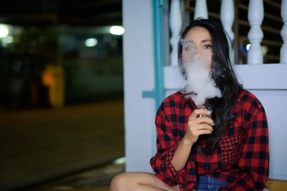 Une femme en chemise à carreaux rouges et noirs exhale un nuage de vapeur d'une cigarette électronique, son souffle se mêlant à la nuit.