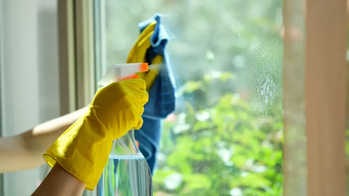 Main tenant, une main gantée de jaune pulvérise du vinaigre blanc sur une fenêtre pour nettoyer la résistance de la saleté.
