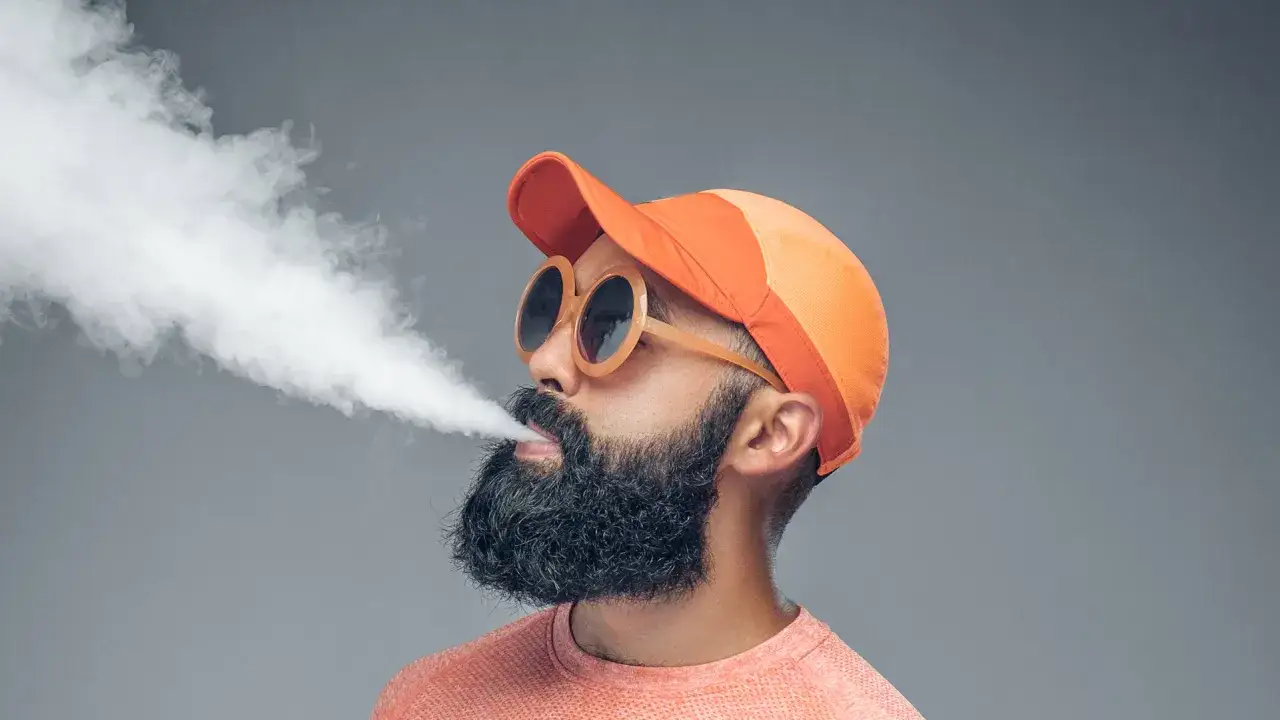 Homme à barbe, casquette orange et lunettes rondes, exhalant un nuage de vapeur de cigarette électronique, peut-être sujet à fatigue et vertiges.