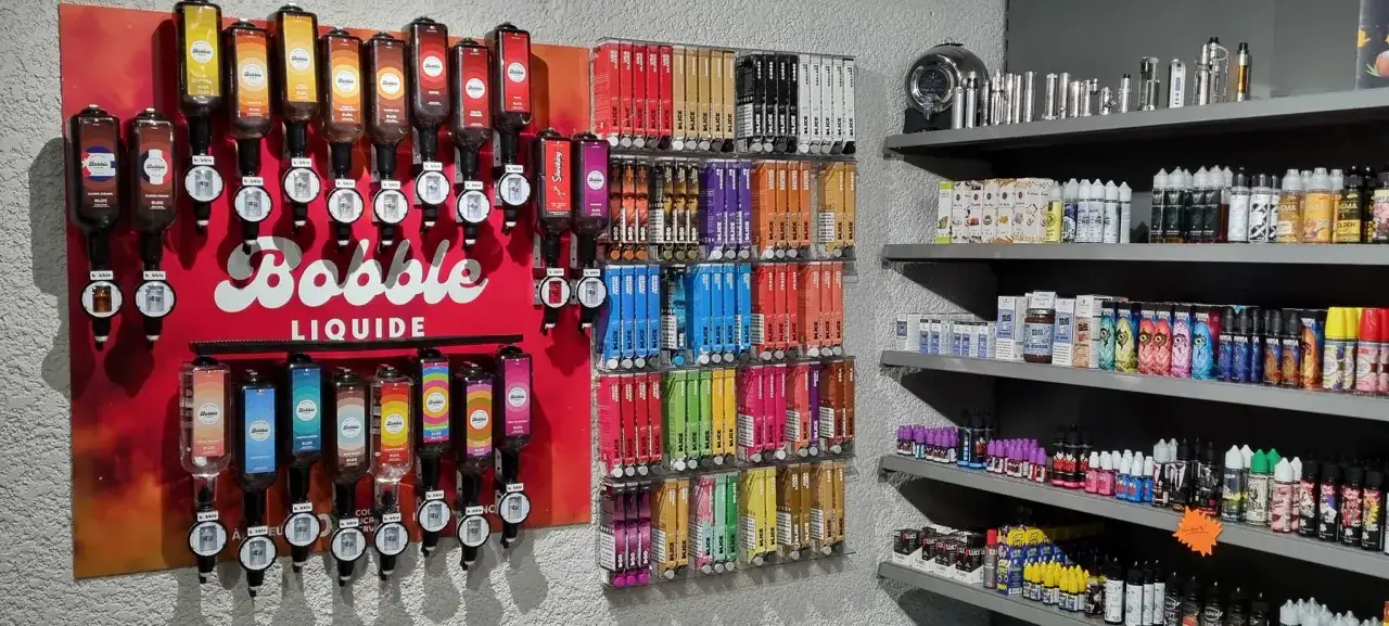 Un mur rempli de distributeurs de bobble e liquide aux couleurs vives, avec des étagères chargées de flacons et de cigarettes électroniques.