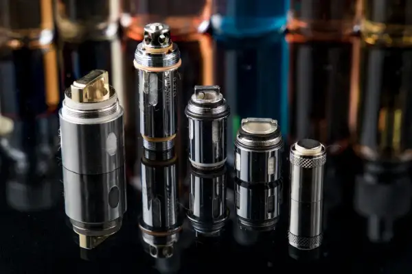 Plusieurs résistances pour cigarette électronique, certaines dorées, d'autres argentées, sont alignées sur un fond sombre avec des flacons de e-liquides flous.