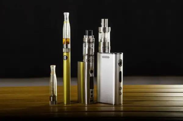 Collection de cigarettes électroniques, dont une dorée et une argentée, sur une table en bois. Ces appareils de vapotage, souvent à basse tension, offrent une alternative moderne.