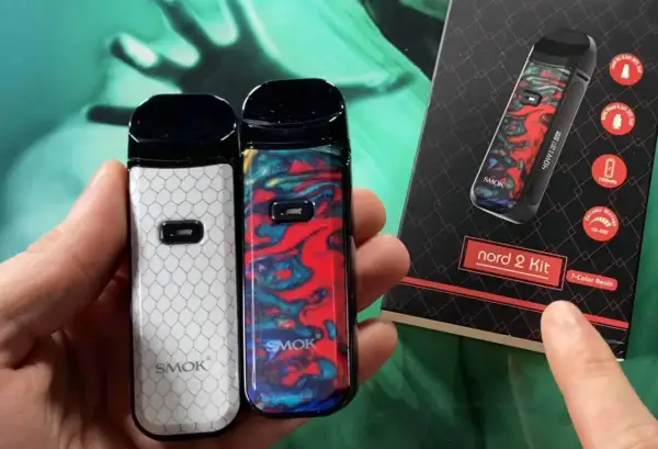 Deux vapoteuses Smok Nord 2, une blanche à motifs nid d'abeille, l'autre colorée. La boîte du kit Smok Nord 2 est visible.
