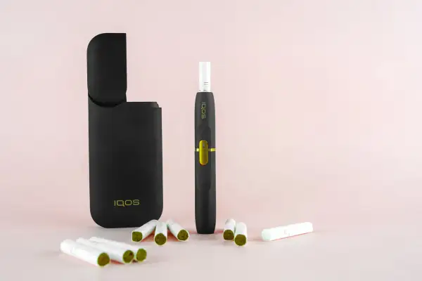 Un étui IQOS noir ouvert, un appareil de cigarette électronique qui chauffe, et plusieurs bâtonnets de tabac sur fond rose pâle.