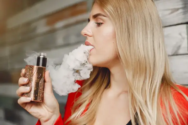 Une femme blonde vapote une cigarette électronique au design unique, exhalant un nuage de vapeur.