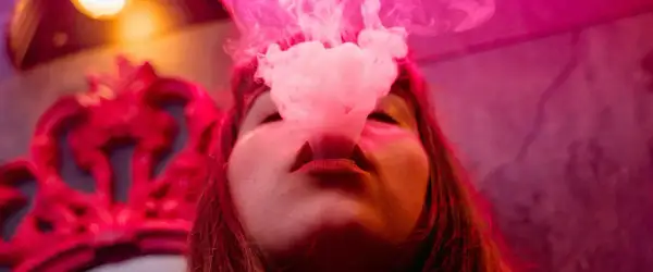 Une femme exhale de la vapeur d'une cigarette électronique, la fumée rose s'élevant autour de son visage. À gauche, une couronne rouge ornée.