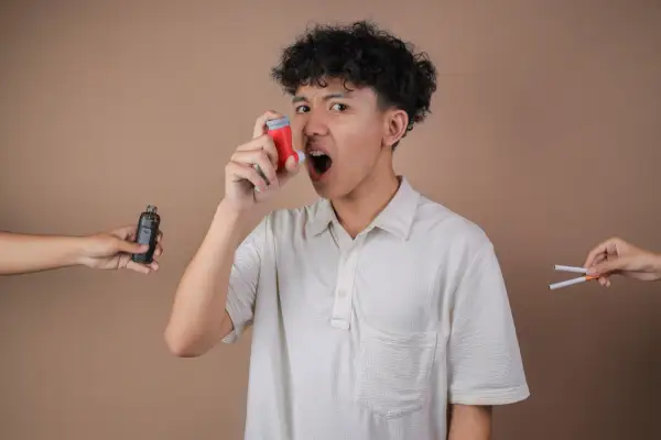 Un jeune homme utilise son inhalateur pour l'asthme, tandis qu'une cigarette électronique et des cigarettes sont présentées de chaque côté, soulignant les dangers.